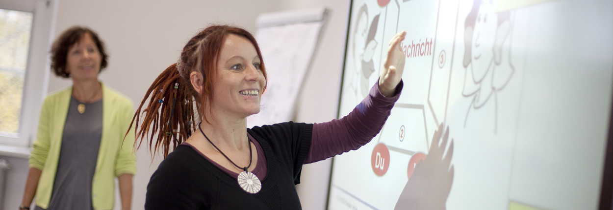 Frau mit Dreadlocks zeigt auf interaktives Whiteboard mit Text 'Schönheit' und Illustrationen, weitere Person im Hintergrund.