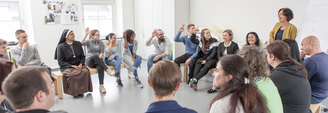 Menschen sitzen im Kreis auf Stühlen in einem hellen Raum, einige heben die Hand, andere hören zu.