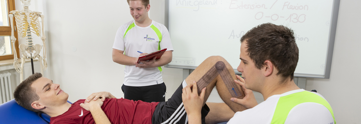 Zwei Männer in Sportkleidung führen eine Beinuntersuchung durch, eine Person steht mit Notizblock vor einem Whiteboard mit medizinischem Text.