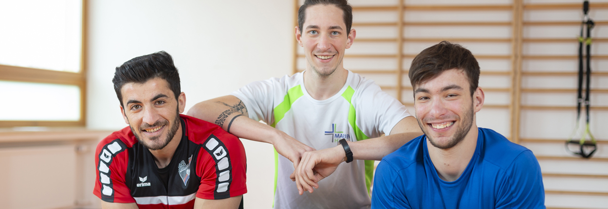 Drei junge Männer in Sportkleidung sitzen nebeneinander, einer trägt ein rotes Trikot, einer ein weißes T-Shirt mit grünem Muster, der dritte ein blaues Shirt.