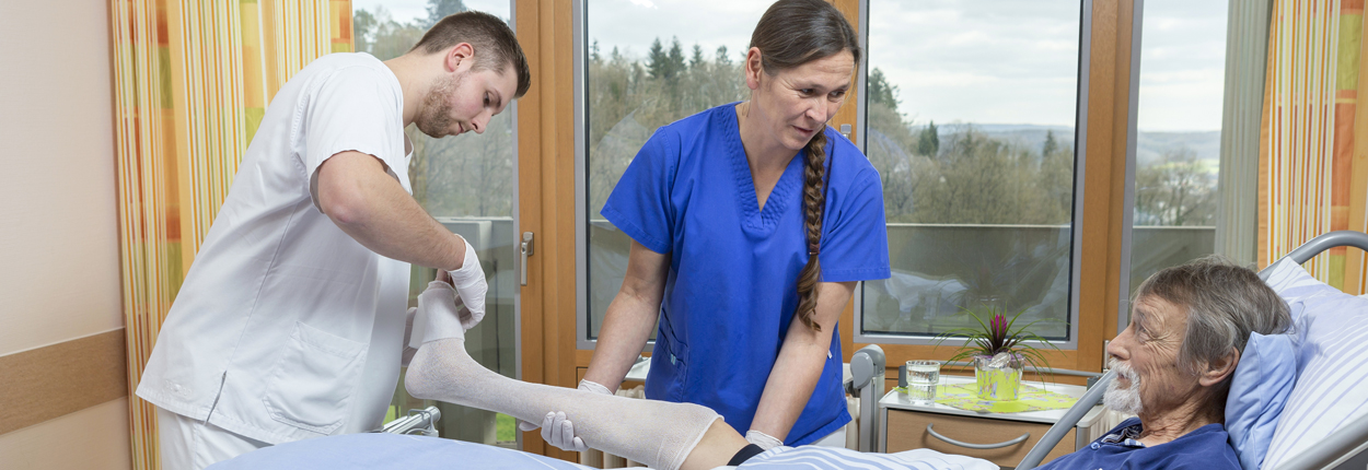 Zwei Pflegekräfte in weißer und blauer Kleidung legen einem liegenden Patienten eine Schiene an einem Bein an, Zimmer mit Fenster und Vorhängen im Hintergrund.