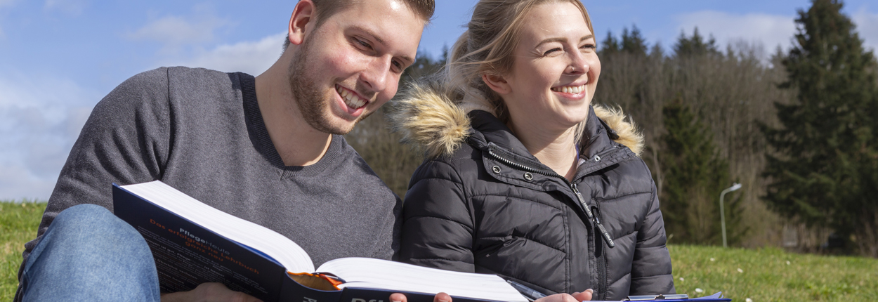 Zwei Personen sitzen auf einer Wiese, eine hält ein aufgeschlagenes Buch, beide tragen Jacken, im Hintergrund Bäume und blauer Himmel.